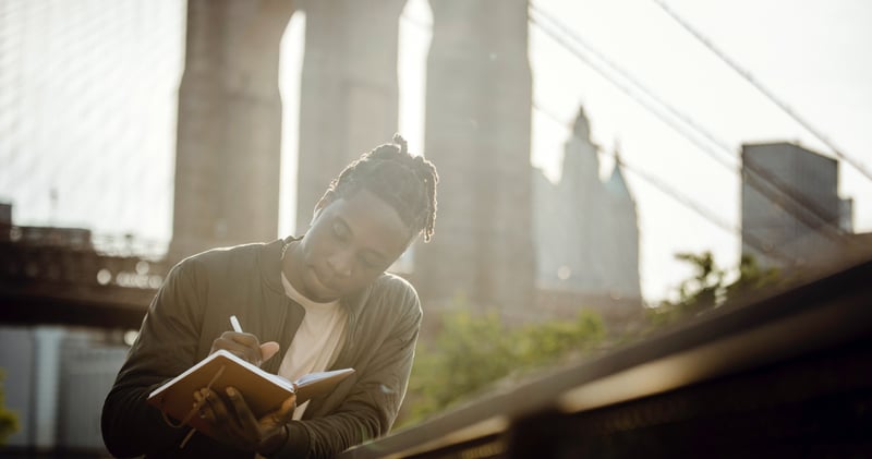 Thoughtful guy writing in notebook leaning on fence against bridge